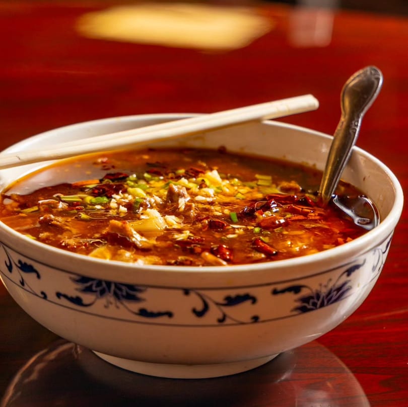 A bowl of spicy Chinese noodle soup with meat, chili flakes, and green onions on a red table.