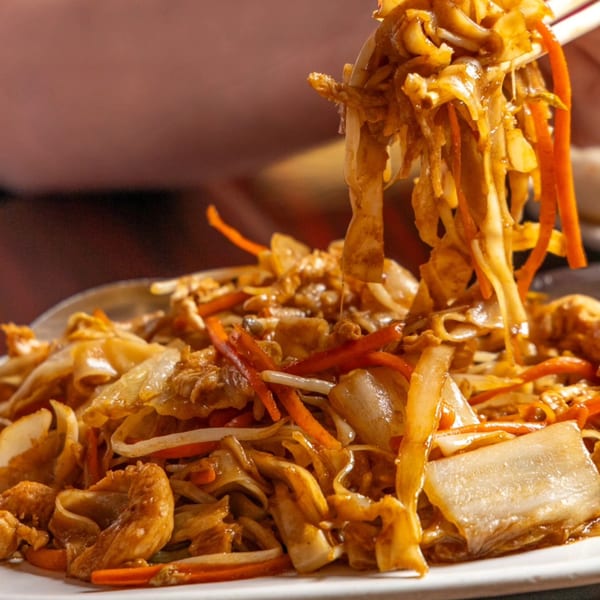 Chopsticks lifting saucy flat noodles with chicken, carrots, and cabbage from a white plate.